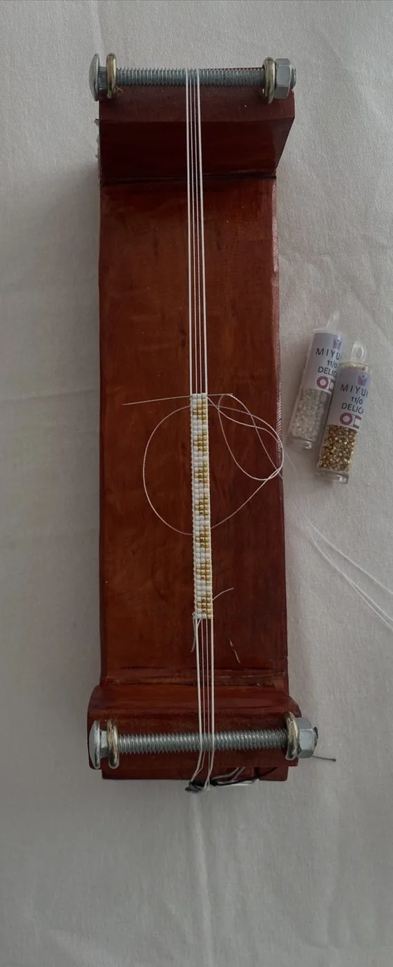 Telar de madera con una pulsera blanca y dorada a medio tejer, junto a dos tubos de mostacillas Miyuki Delica